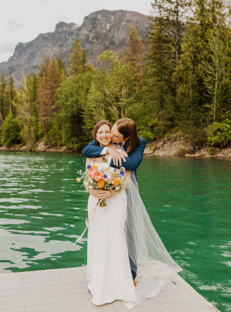 Groom kissing bride on dock on Lake McDonald in Glacier National Park. Bright floral wedding bouquet.