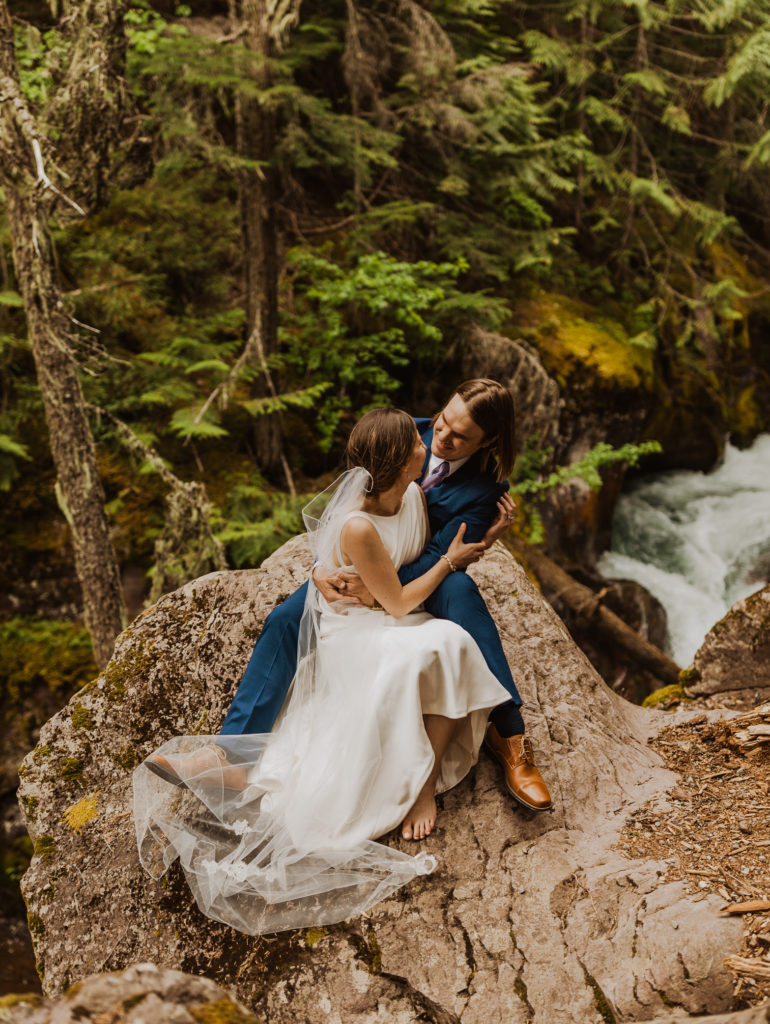 Bride and groom in Glacier National Park sitting together above river after Lake McDonald Elopement