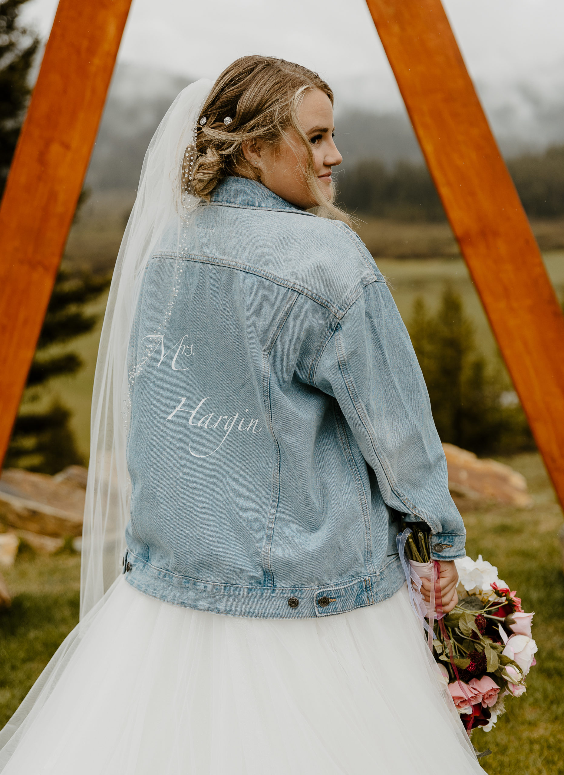 Montana bride with denim jacket, veil and wedding bouquet standing under a triangle arch