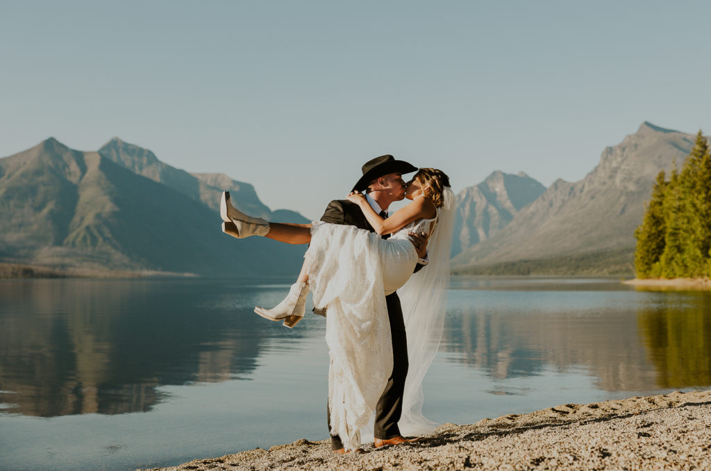 Groom carrying bride in front of Lake McDonald. Glacier National Park Photography by Haley Jessat
