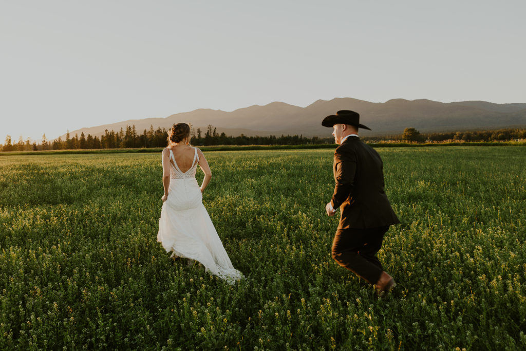 Bride and groom with hats in a grass field and mountains behind them. Glacier National Park Photography by Haley Jessat