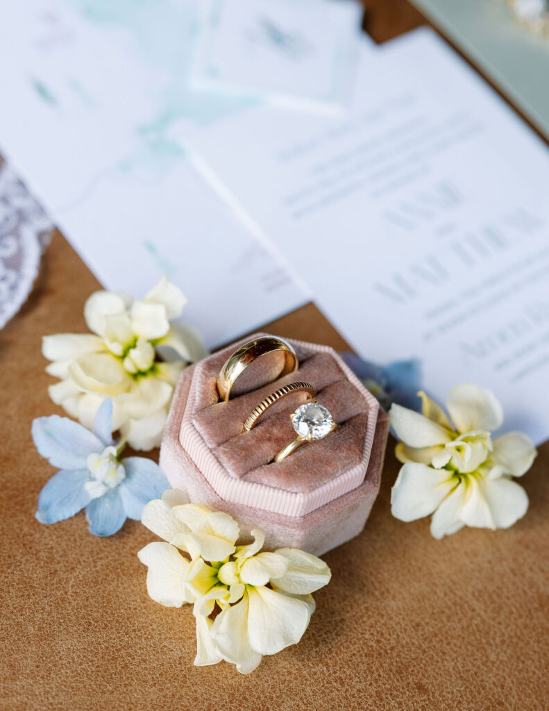 Close-up of wedding rings in a velvet ring box, surrounded by pastel flowers, with wedding invitations in the background, showcasing the wedding details at a Montana wedding.