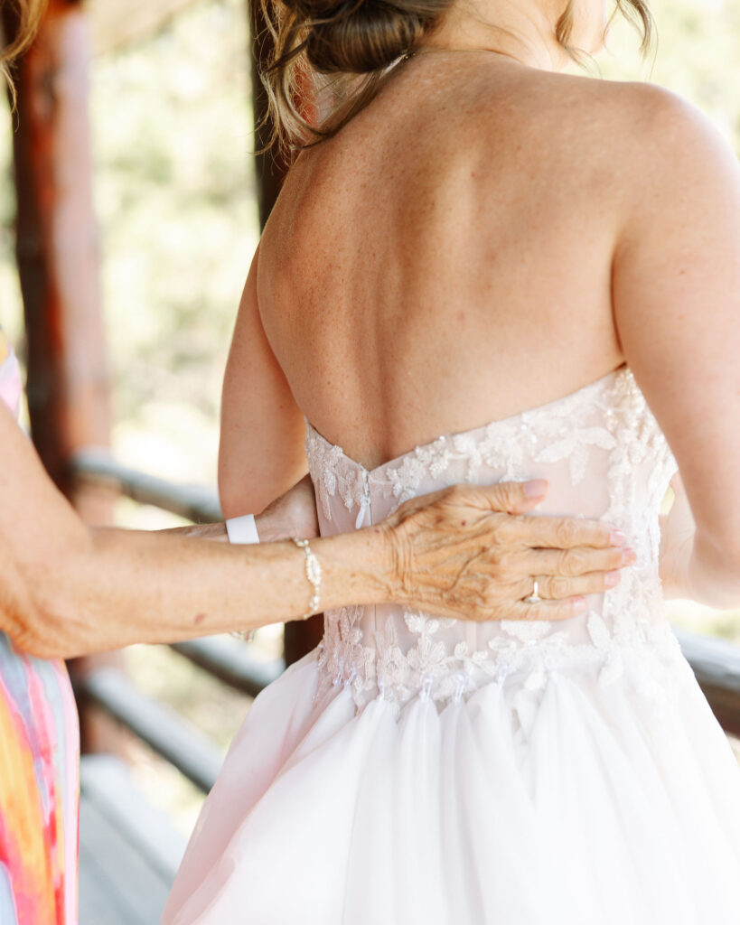 Close-up of a bride’s back in a lace wedding dress, with an elderly woman’s hand gently touching her, capturing a tender pre-ceremony moment at Amen Ranch.