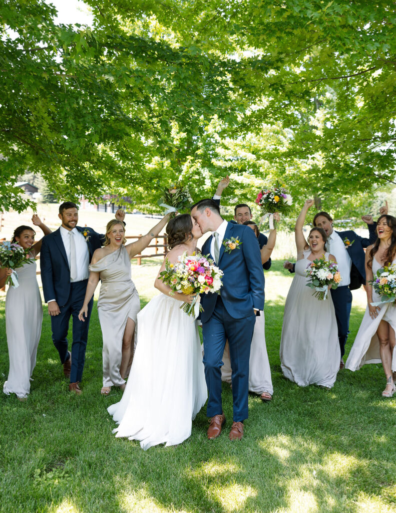 Bride and groom joyfully walking with their bridal party under a large tree, celebrating their wedding day at Amen Ranch in Montana.