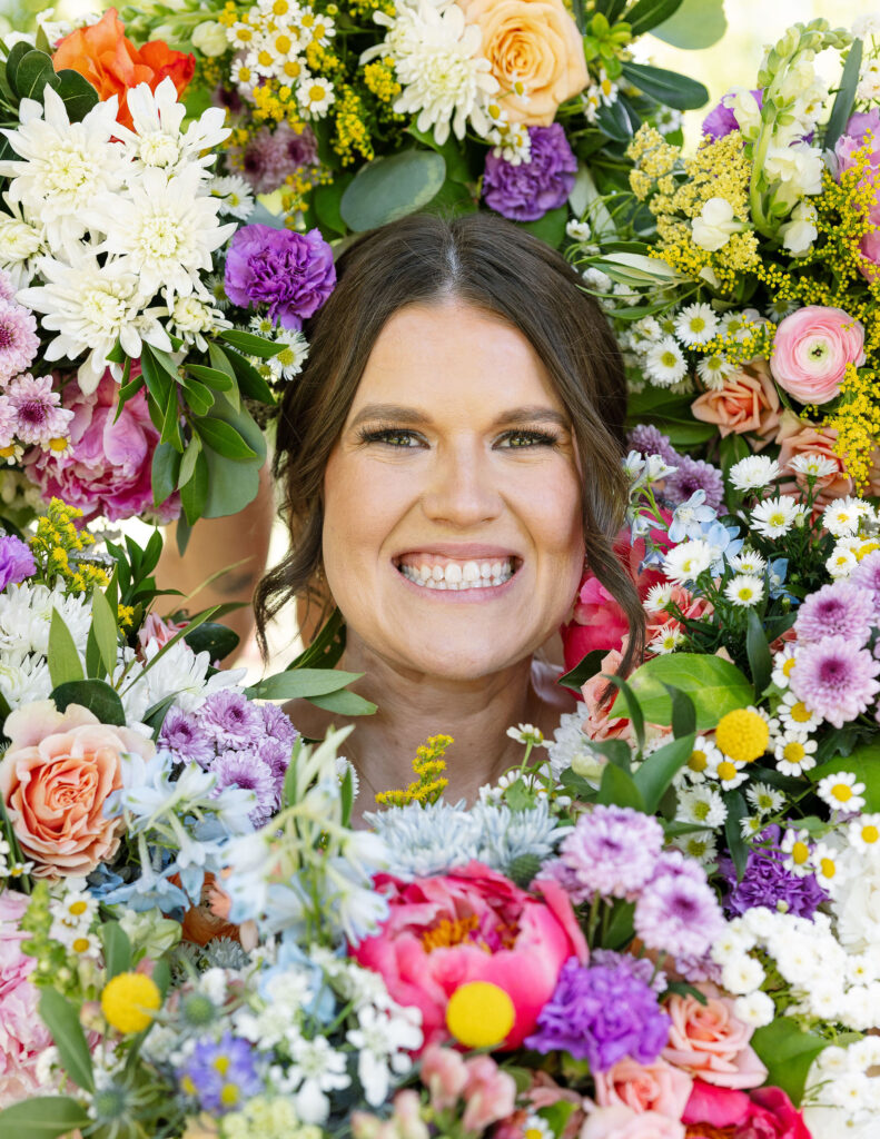 Bride smiling brightly while holding a colorful, vibrant bouquet close to her face, showcasing the floral beauty of a Montana wedding.

