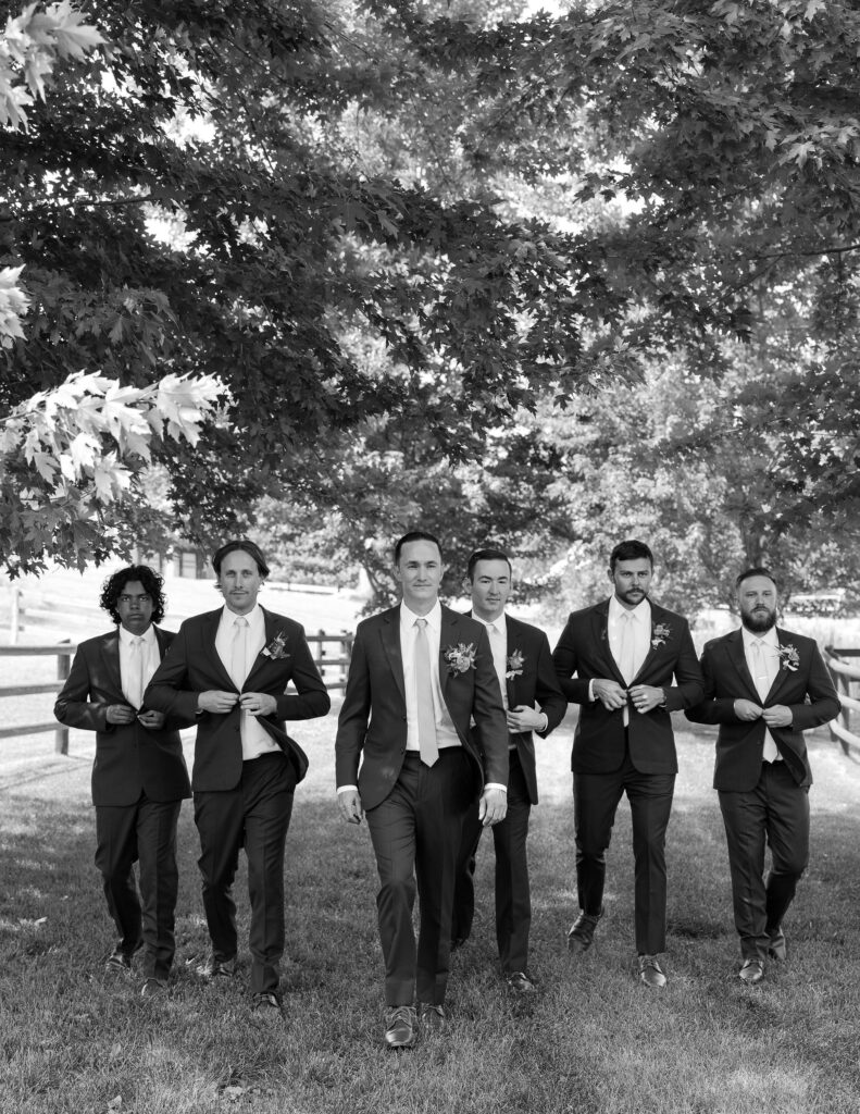 Black and white image of the groom and groomsmen walking confidently under shaded trees, dressed in formal suits at a Montana ranch wedding venue.