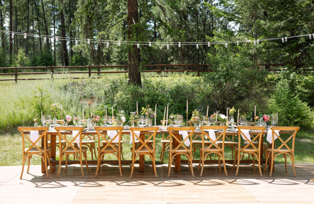 Outdoor reception area with rustic wooden tables and string lights, set amidst tall trees, creating a romantic atmosphere at a Montana ranch wedding.