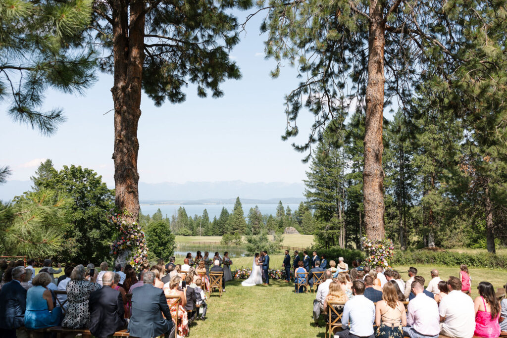 Wide shot of a wedding ceremony under tall pine trees with guests seated, capturing the natural beauty and intimate setting of a Montana wedding venue.