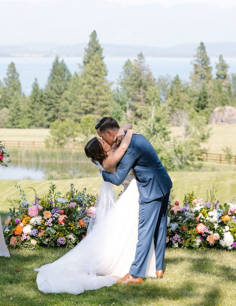 Bride and groom sharing a joyful embrace and kiss after being pronounced married, surrounded by vibrant floral arrangements and a stunning natural backdrop at Amen Ranch in Montana.