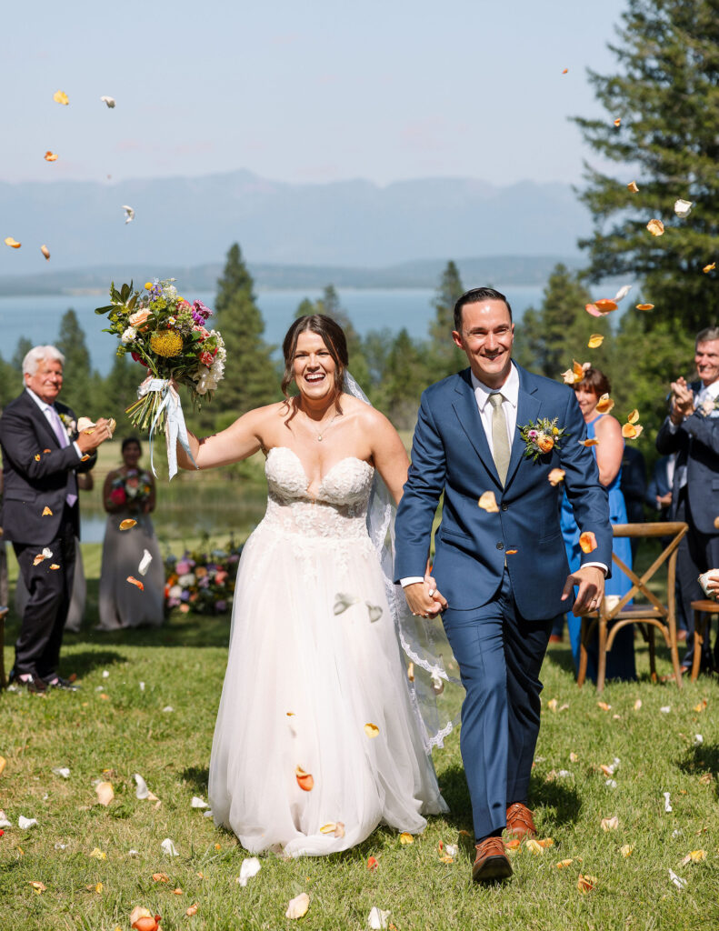 Bride and groom smiling and holding hands as they walk down the aisle, with flower petals in the air, celebrating their newlywed status at a Montana ranch wedding.