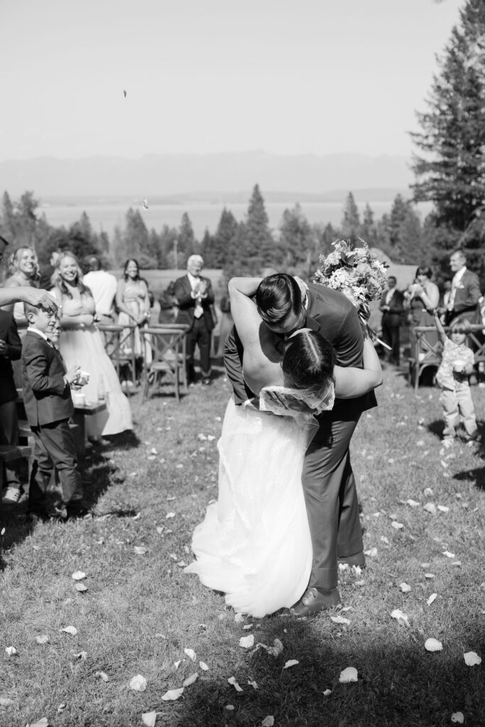 Black and white image of the groom dipping the bride for a kiss during their recessional walk, surrounded by cheering guests and flower petals at Amen Ranch.