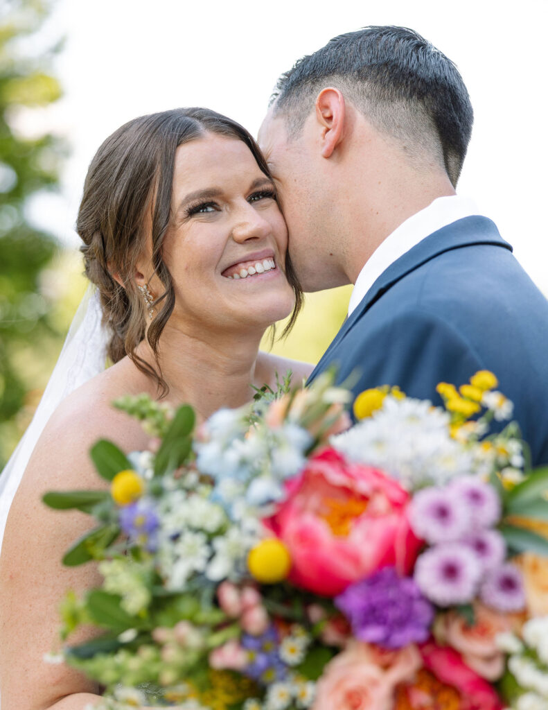 Bride smiling joyfully while the groom kisses her cheek, holding a vibrant bouquet, capturing a loving moment at Amen Ranch.