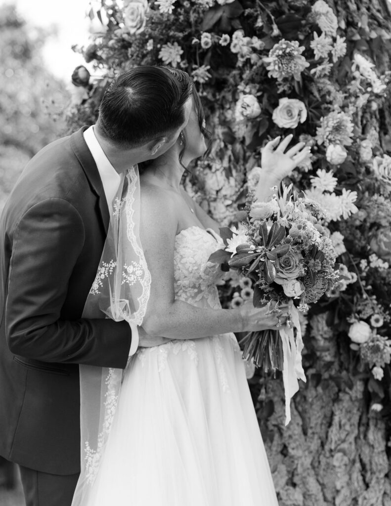 Black and white close-up of the bride and groom sharing a kiss in front of a floral backdrop, capturing an intimate and romantic moment at a Montana ranch wedding.