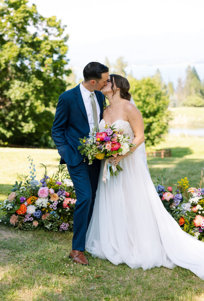 Bride and groom kissing while holding a vibrant bouquet, standing in front of colorful floral arrangements, showcasing a romantic moment at Amen Ranch in Montana.