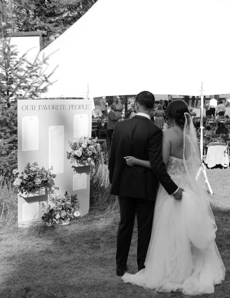 Black and white photo of the bride and groom standing arm in arm, looking at the "Our Favorite People" seating chart display, capturing a thoughtful moment at Amen Ranch in Montana.