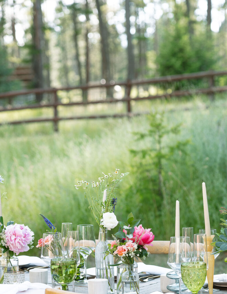 Elegant wedding table setup with glassware, candles, and floral centerpieces, featuring a mix of pink and white flowers, set against a lush green backdrop at a Montana ranch wedding.