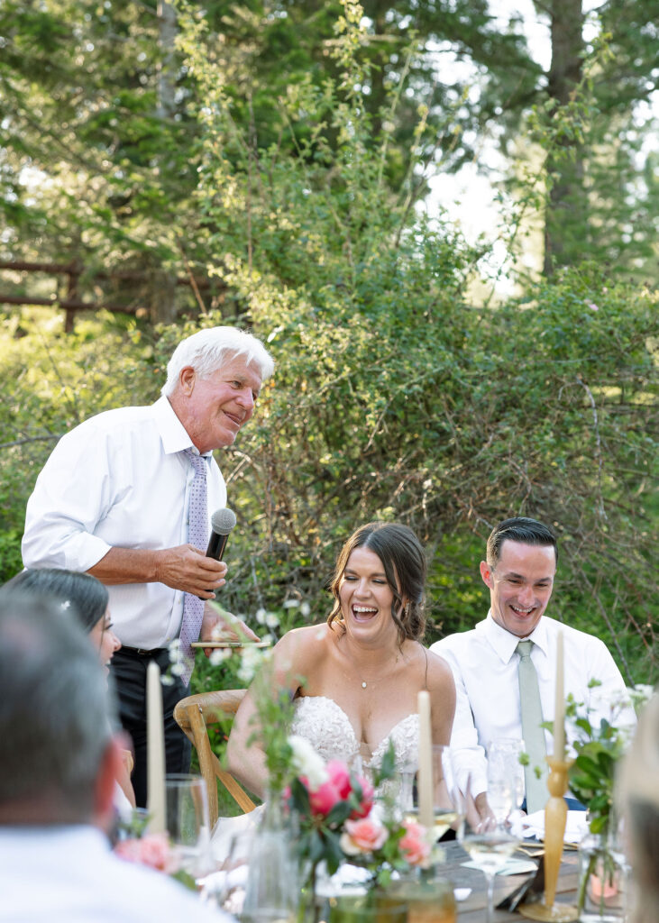 Father of the bride giving a heartfelt speech with a microphone, while the bride and groom smile and listen attentively at their wedding reception at Amen Ranch.