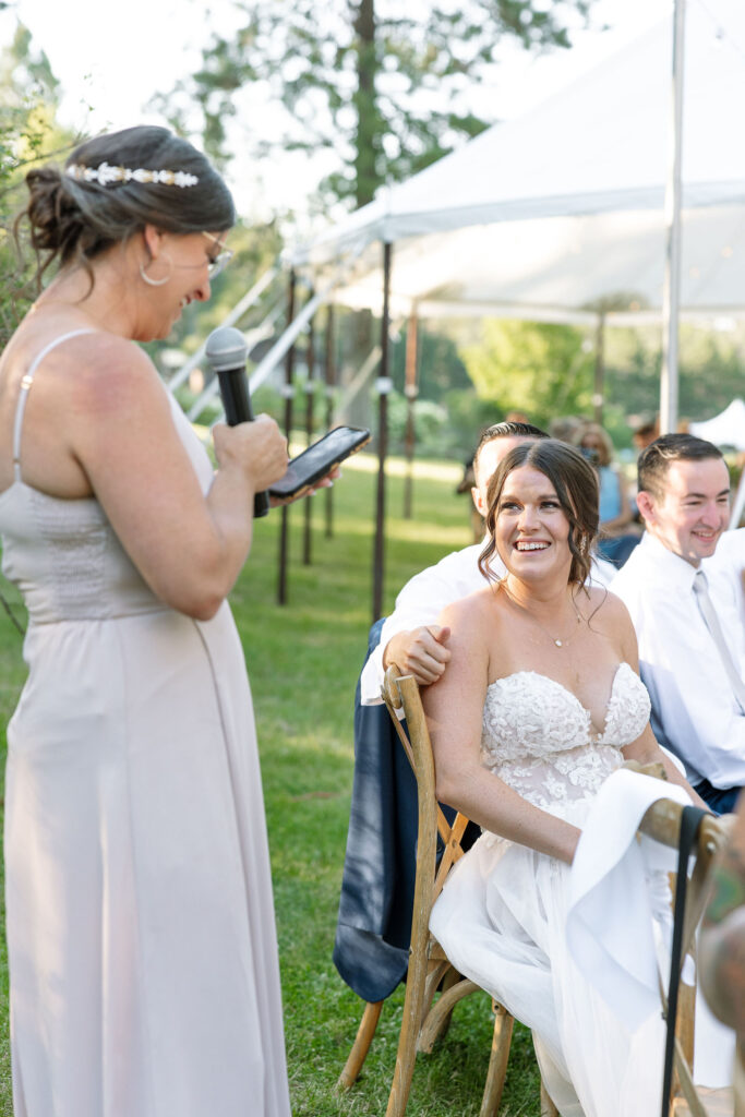 Bridesmaid giving a touching speech with a microphone while the bride sits and smiles, enjoying the heartfelt words during the reception at Amen Ranch.