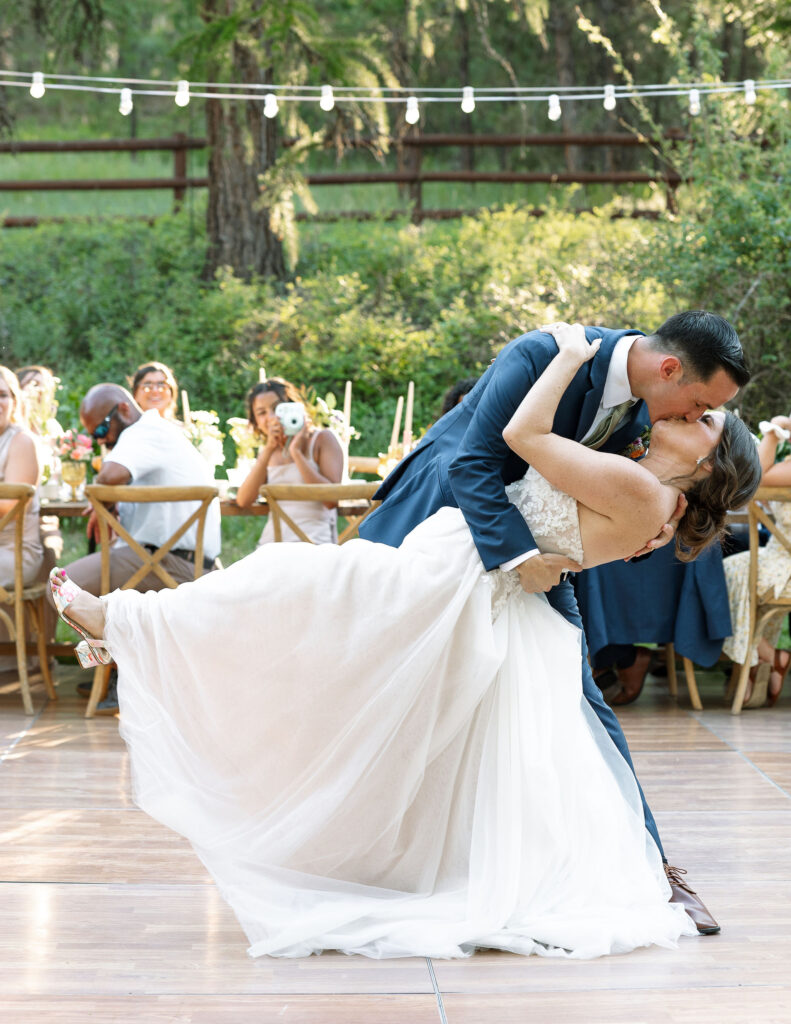 Groom dipping the bride for a romantic kiss during their first dance, surrounded by guests under string lights at a Montana ranch wedding.