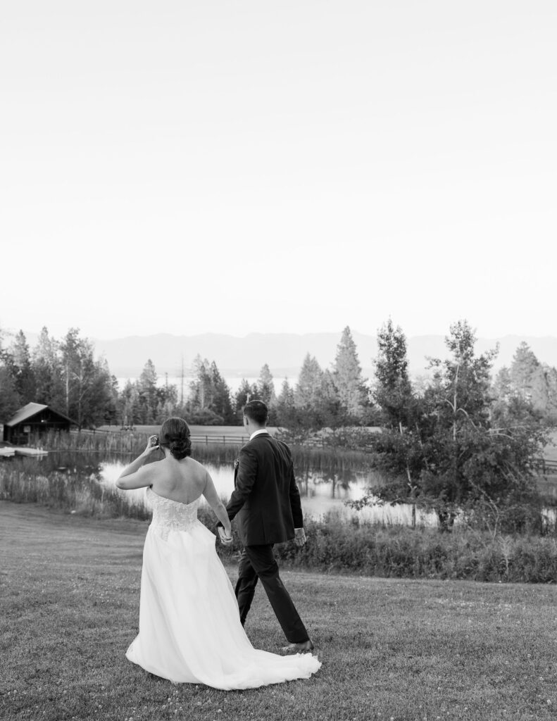 Black and white photo of the bride and groom walking hand in hand across a grassy field, with a scenic mountain backdrop at Amen Ranch in Montana.