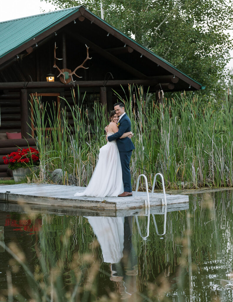 Bride and groom embracing on a wooden dock in front of a rustic cabin at Amen Ranch, reflecting in the serene pond, highlighting a romantic Montana wedding venue.