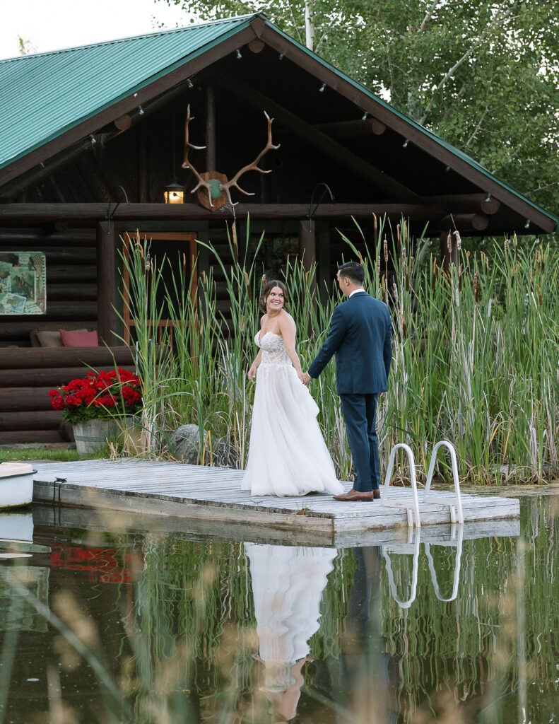 Bride and groom holding hands on a wooden dock in front of a rustic cabin at Amen Ranch, a serene Montana wedding venue.