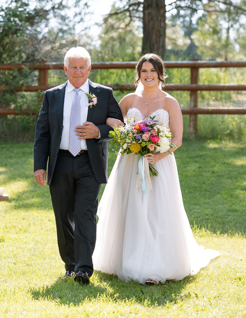 Bride walking down the aisle with her father, smiling and holding a vibrant bouquet, capturing a touching moment at a Montana ranch wedding.