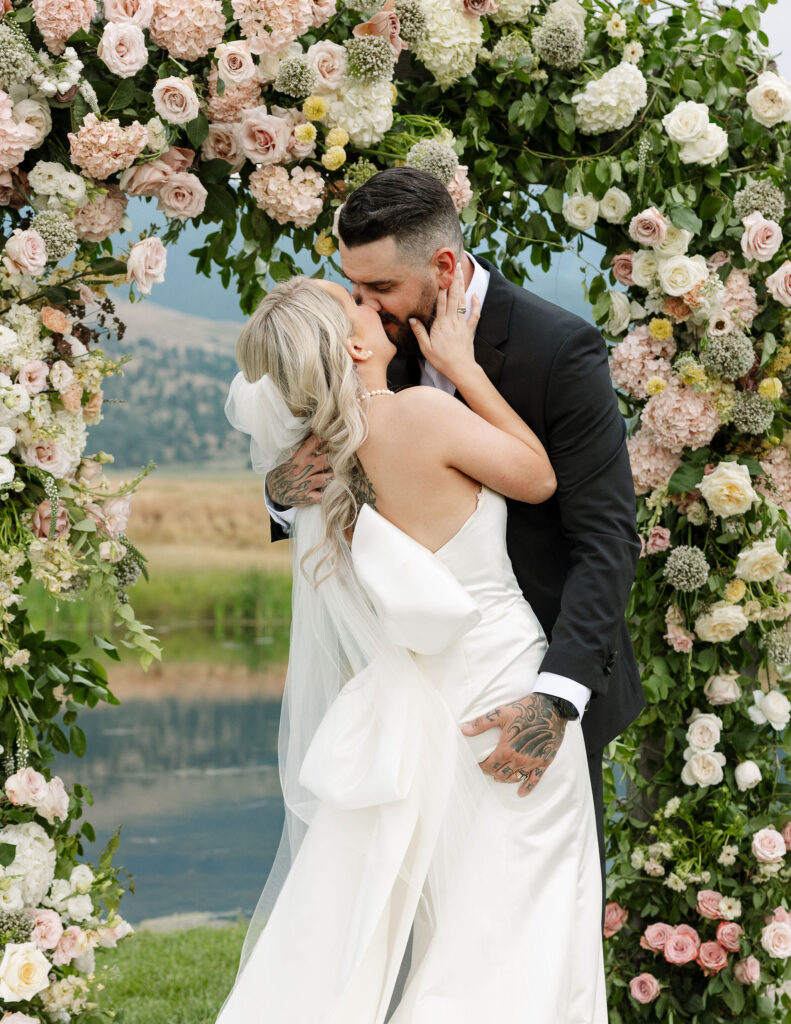 The bride and groom share a kiss under a floral arch during their outdoor ceremony at Copper Rose Ranch Wedding Venue. Captured by Haley J Photo, a Montana Wedding Venue Photographer.