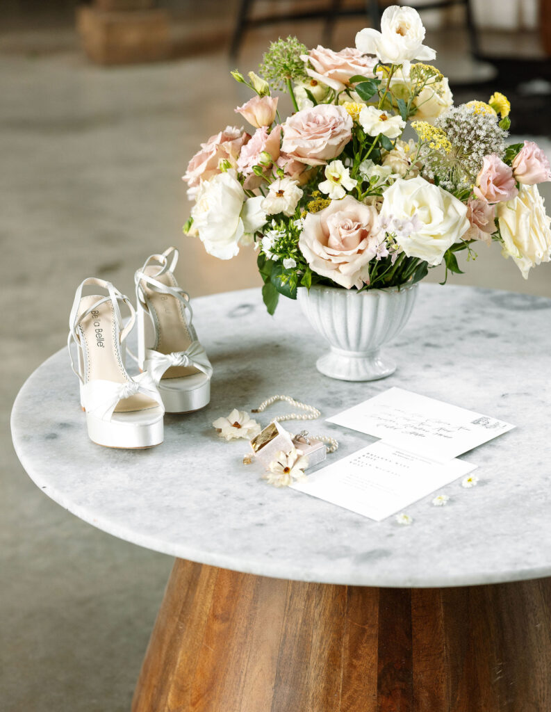 Wedding details, including shoes, invitations, and a floral arrangement, displayed on a round marble table at Copper Rose Ranch Wedding Venue. Captured by Haley J Photo, a Montana Wedding Venue Photographer.