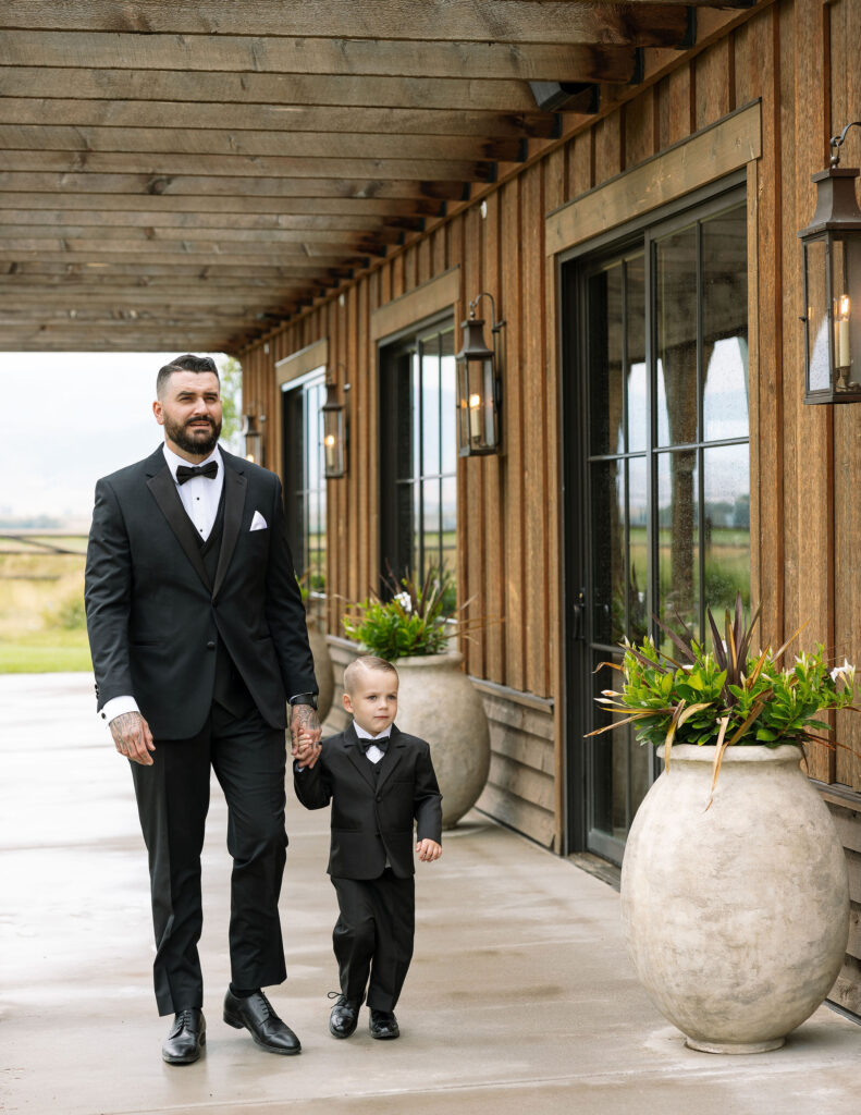 The groom walking hand in hand with a young boy, both dressed in black suits, along the porch of Copper Rose Ranch Wedding Venue. Captured by Montana Wedding Photographer Haley J Photo.