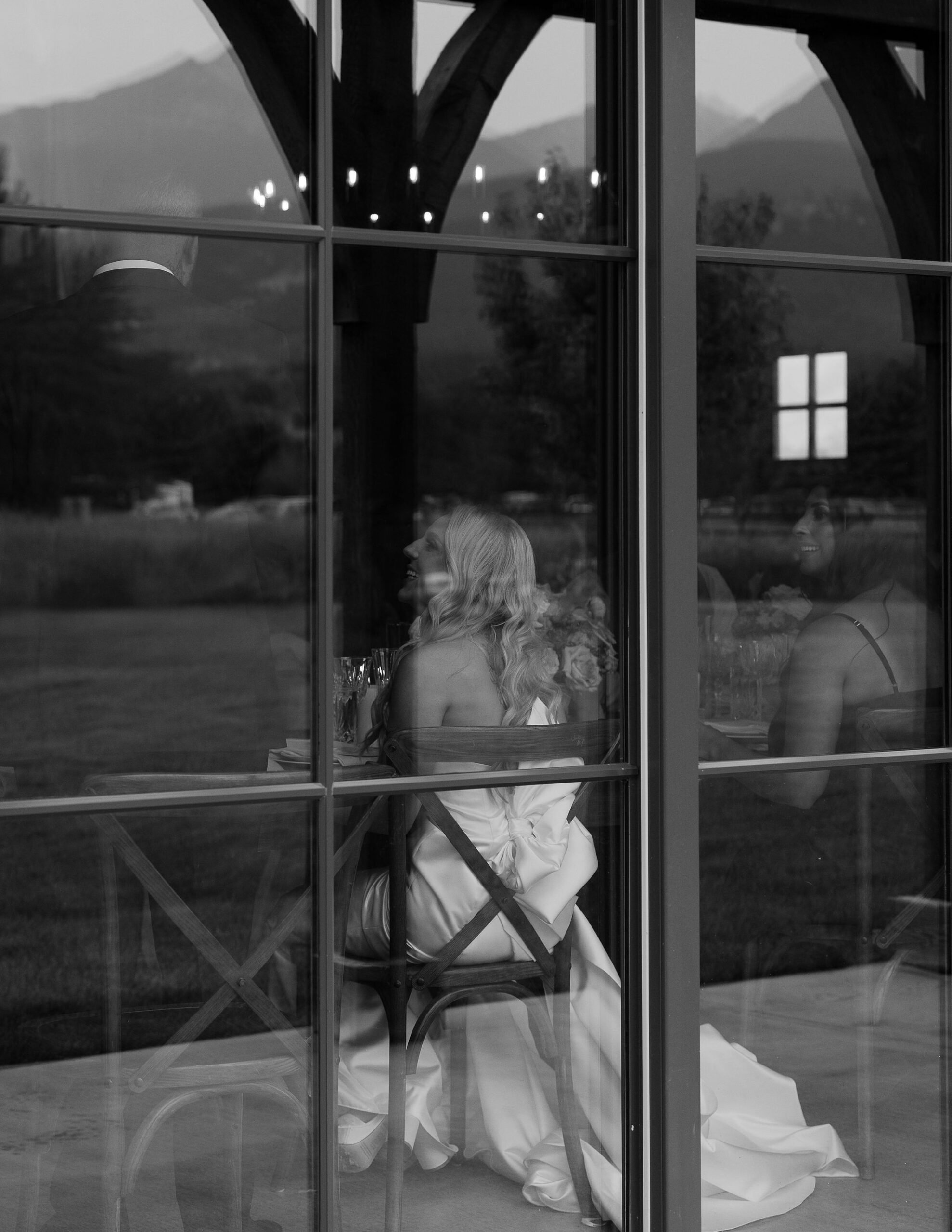  A black and white image of the bride, seated and seen through the window panes of Copper Rose Ranch Wedding Venue. The Montana Bride appears reflective and serene, captured by Montana Wedding Venue Photographer Haley J Photo.