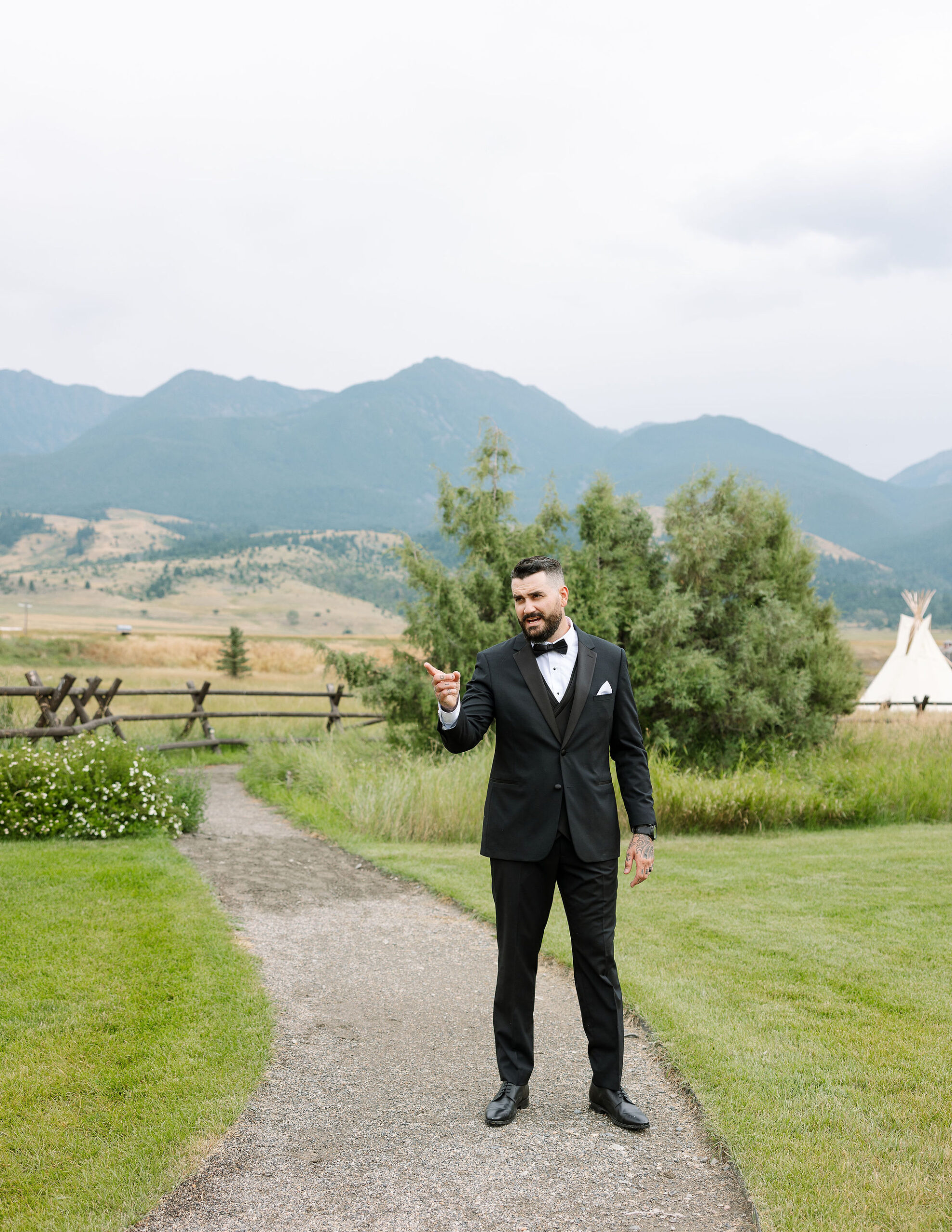 The groom, dressed in a black tuxedo, stands on a path at Copper Rose Ranch Wedding Venue, with the Montana mountains behind him. He is captured in a candid moment by Haley J Photo, a Montana Wedding Venue Photographer.