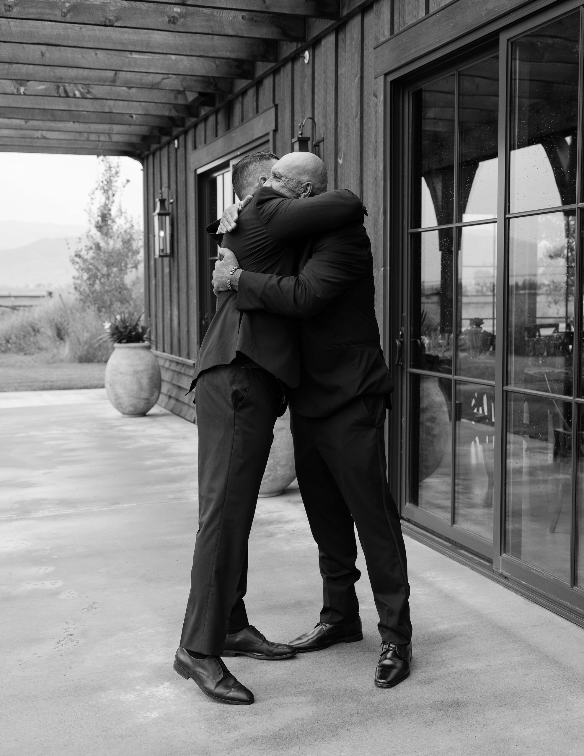 Two men embrace in a warm hug on the porch of the rustic venue at Copper Rose Ranch in Montana. The emotional moment between friends or family members is captured by Montana Wedding Photographer Haley J Photo.