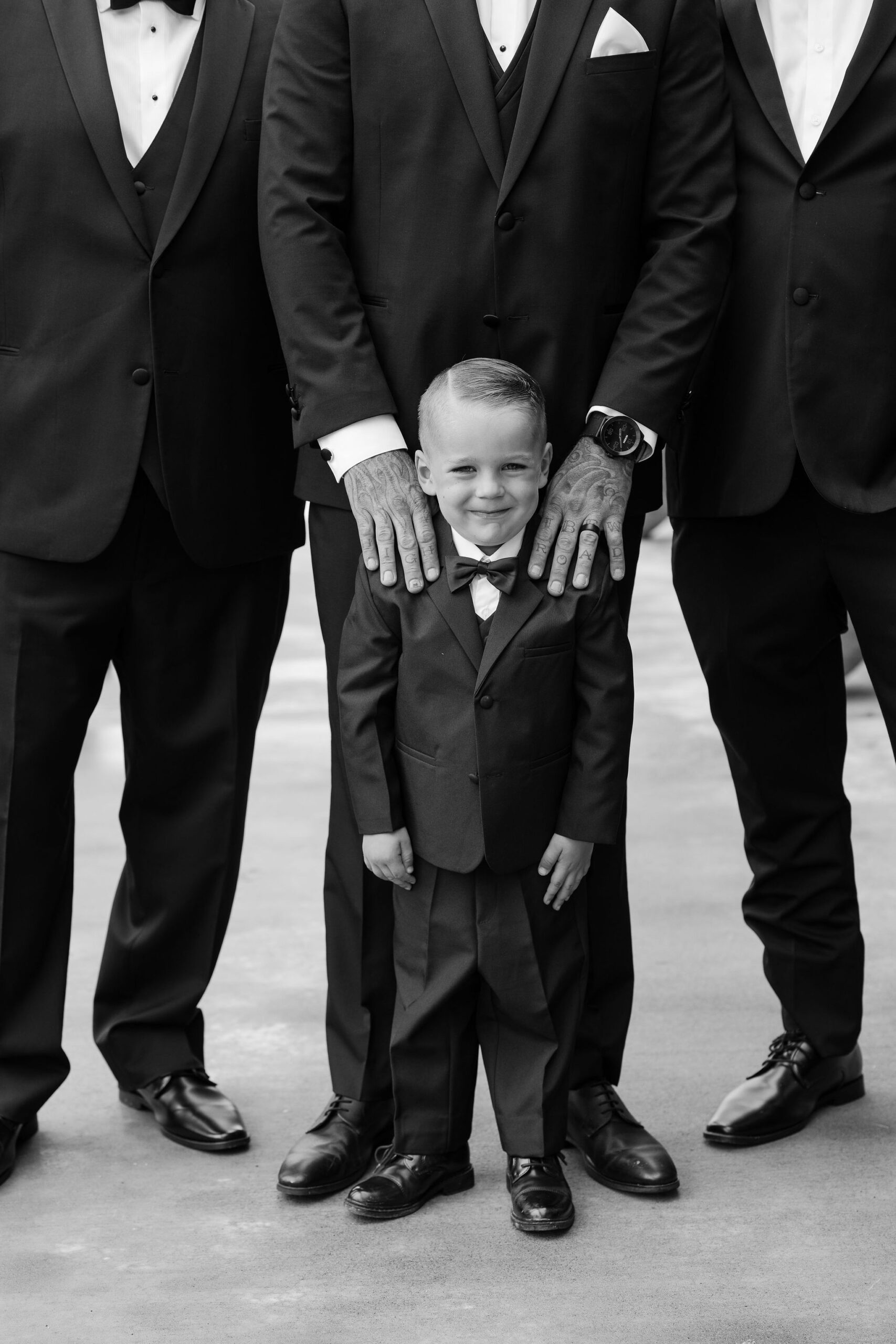 A black and white image of a young boy, smiling proudly, standing in front of the groomsmen dressed in black suits at Copper Rose Ranch Wedding Venue. The playful moment is captured by Montana Wedding Photographer Haley J Photo.