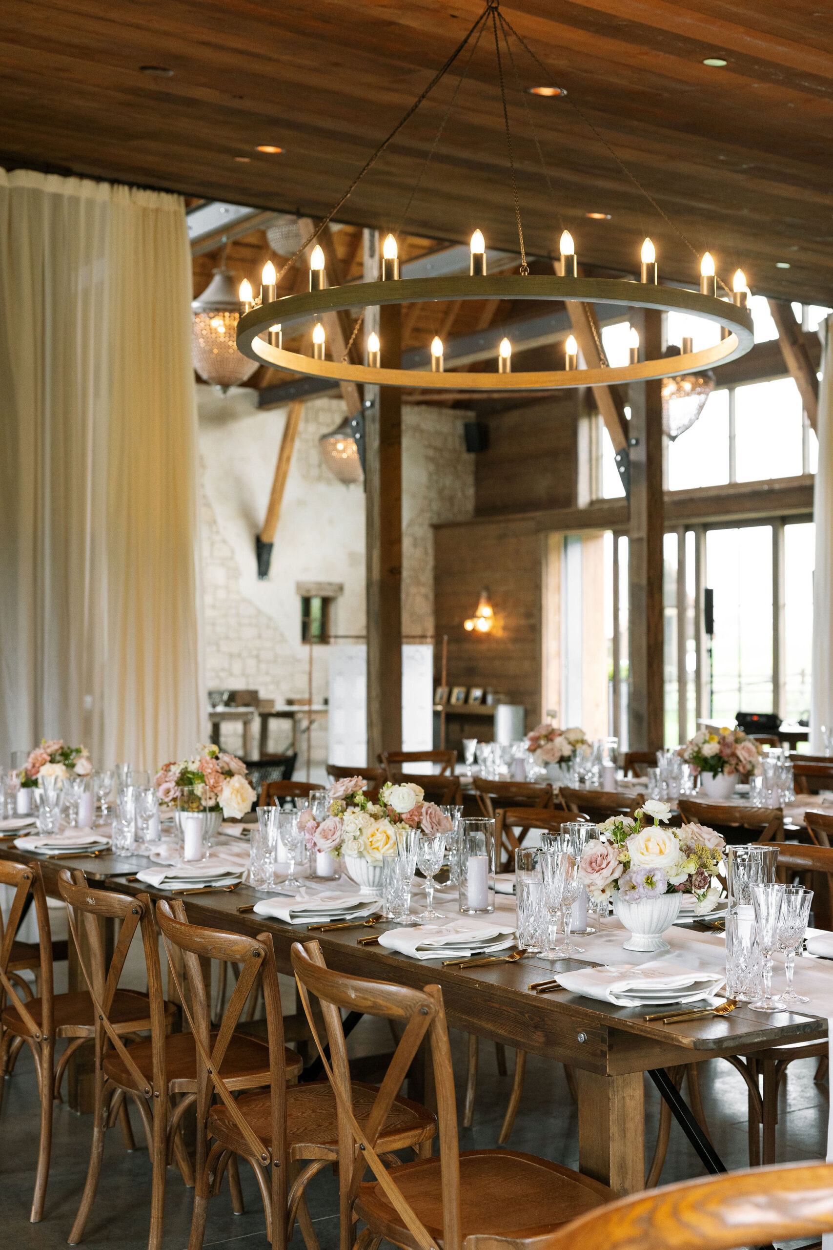 The reception area at Copper Rose Ranch Wedding Venue is set with long wooden tables, cross-back chairs, and soft floral centerpieces. The warm, inviting atmosphere is enhanced by the rustic chandeliers, captured by Haley J Photo, a Montana Wedding Venue Photographer.