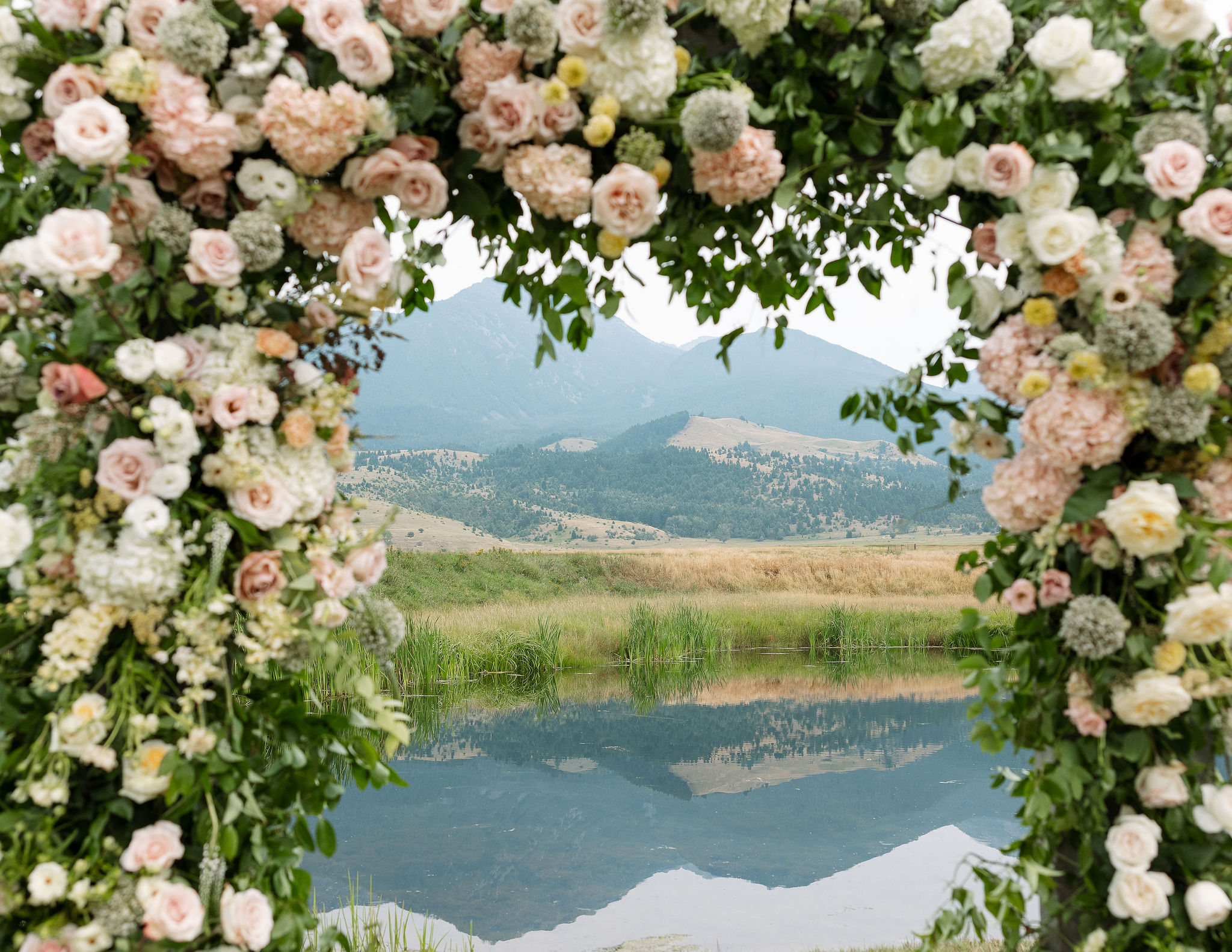 A beautifully decorated floral arch with soft pink and white roses frames a serene view of the pond and mountains at Copper Rose Ranch Wedding Venue in Montana. The reflection of the mountains in the water enhances the natural beauty of the venue, captured by Haley J Photo, a Montana Wedding Venue Photographer.