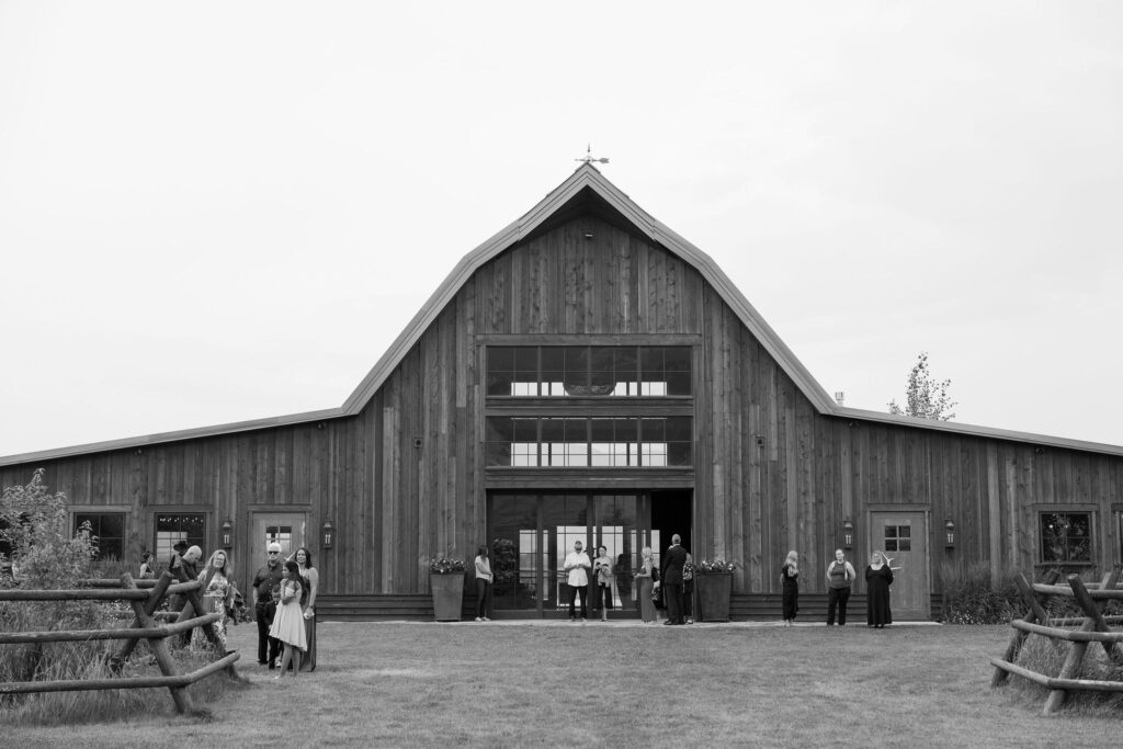 A black and white image of guests gathering outside the barn at Copper Rose Ranch Wedding Venue. Captured by Haley J Photo, a Montana Wedding Venue Photographer.