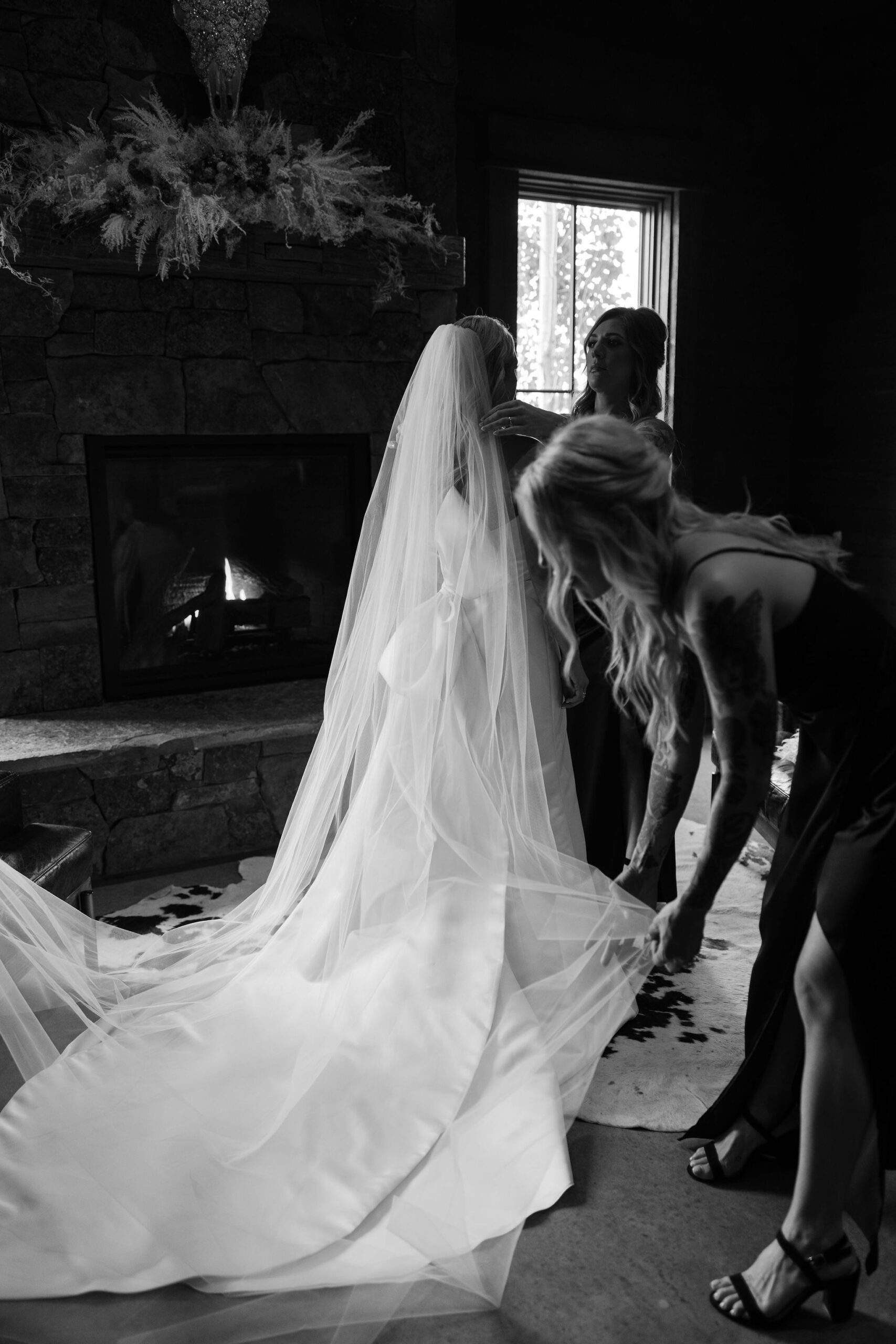 The bride, with a long veil cascading down, is being attended to by her bridesmaids indoors at Copper Rose Ranch Wedding Venue. The black and white image captures the quiet preparation before the ceremony, photographed by Montana Wedding Photographer Haley J Photo.