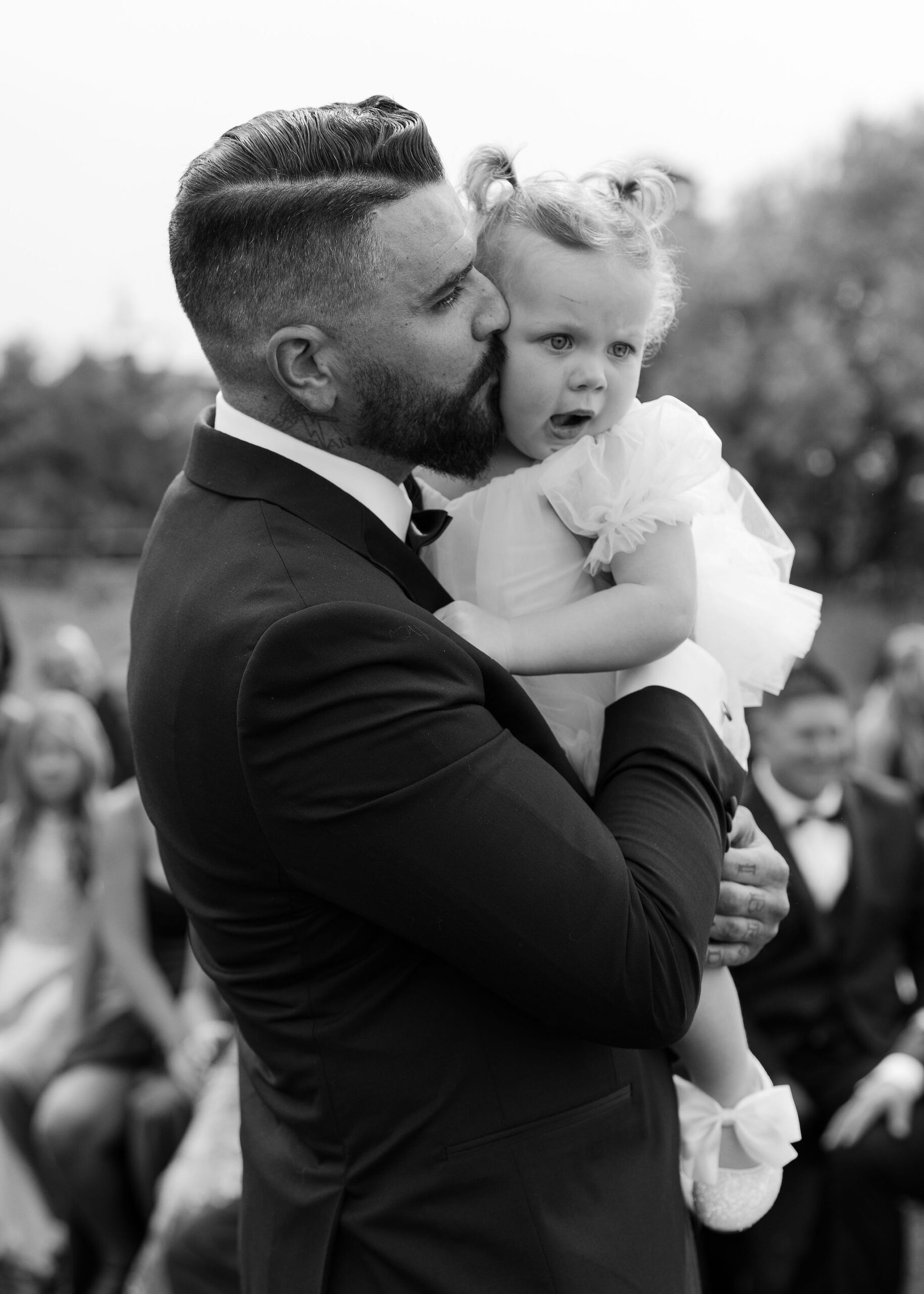 The groom, dressed in a black tuxedo, tenderly kisses a young flower girl on the cheek at Copper Rose Ranch Wedding Venue. The black and white image captures the affectionate moment, photographed by Montana Wedding Photographer Haley J Photo.