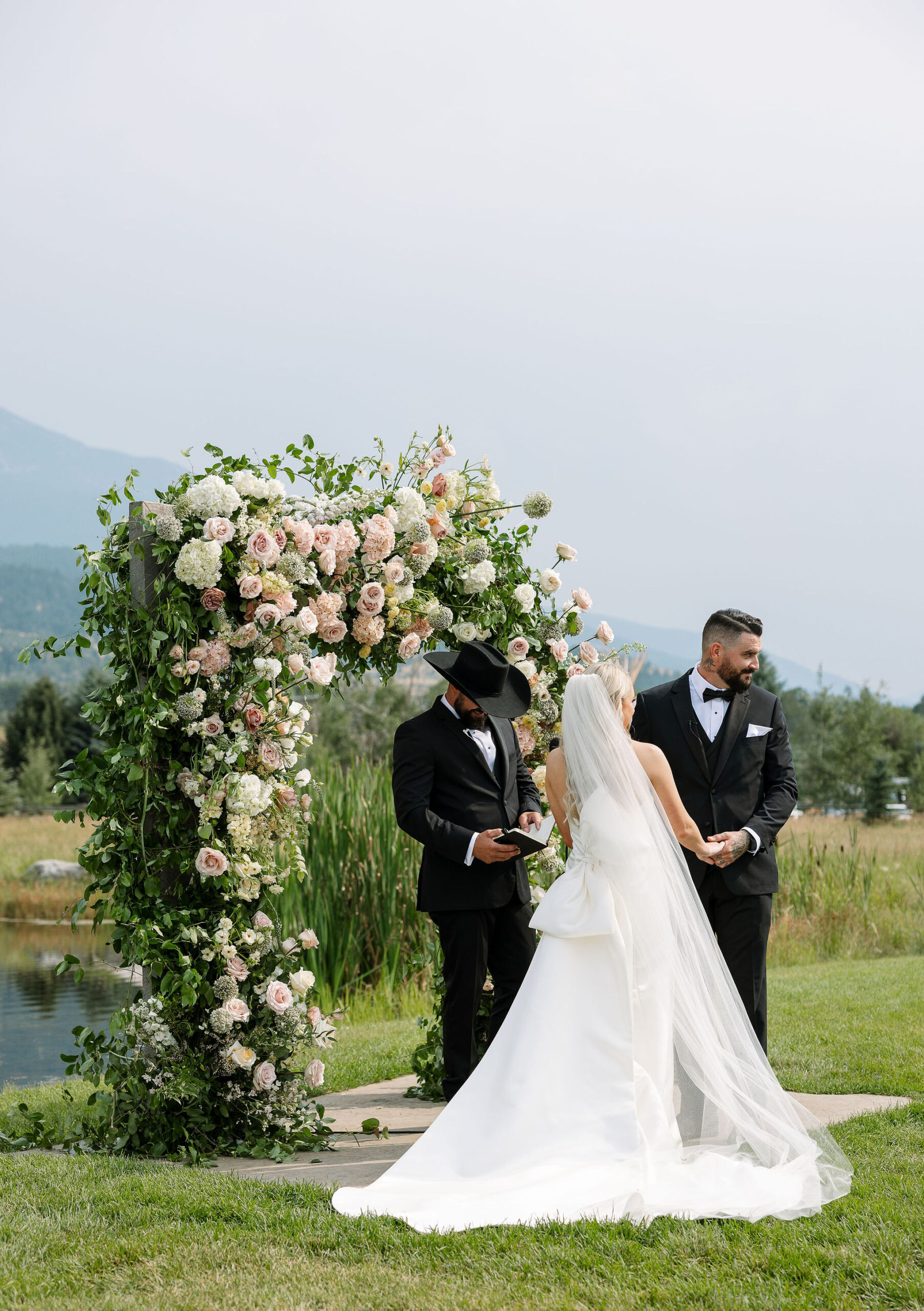 The bride and groom stand under a floral arch during their outdoor ceremony at Copper Rose Ranch Wedding Venue in Montana. The lush greenery and stunning mountain views set the scene for this special moment, captured by Haley J Photo, a Montana Wedding Venue Photographer.