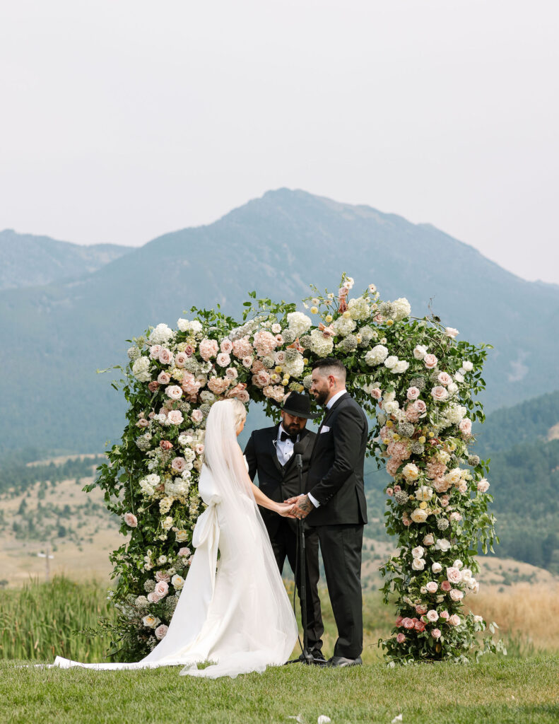 The bride and groom standing under a floral arch during their outdoor ceremony at Copper Rose Ranch Wedding Venue, with Montana mountains as the backdrop. Captured by Haley J Photo, a Montana Wedding Venue Photographer.