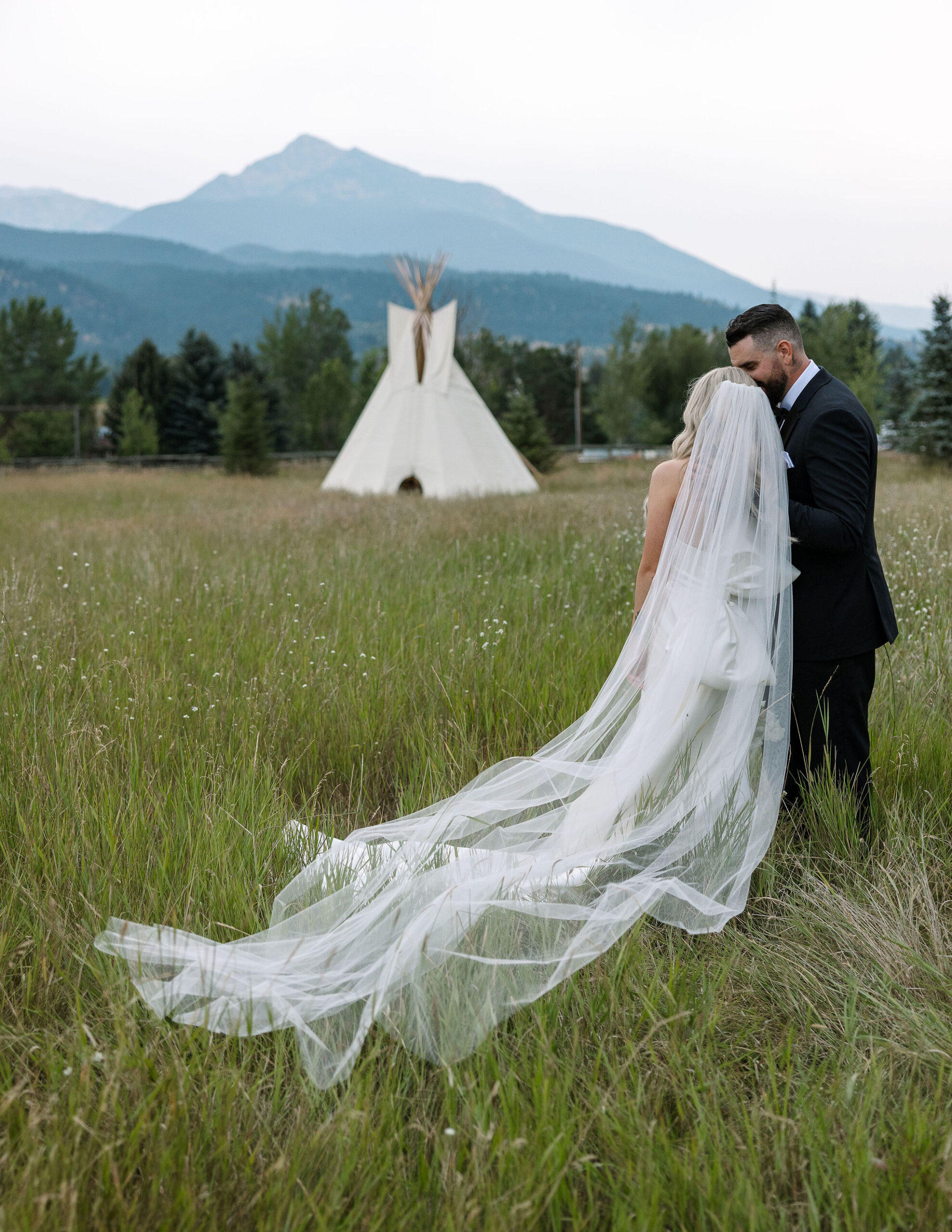 The bride and groom walk hand in hand through a meadow with a traditional teepee and mountains in the background at Copper Rose Ranch Wedding Venue in Montana. The Montana Bride’s long veil trails behind her, and the moment is beautifully captured by Haley J Photo, a Montana Wedding Venue Photographer.