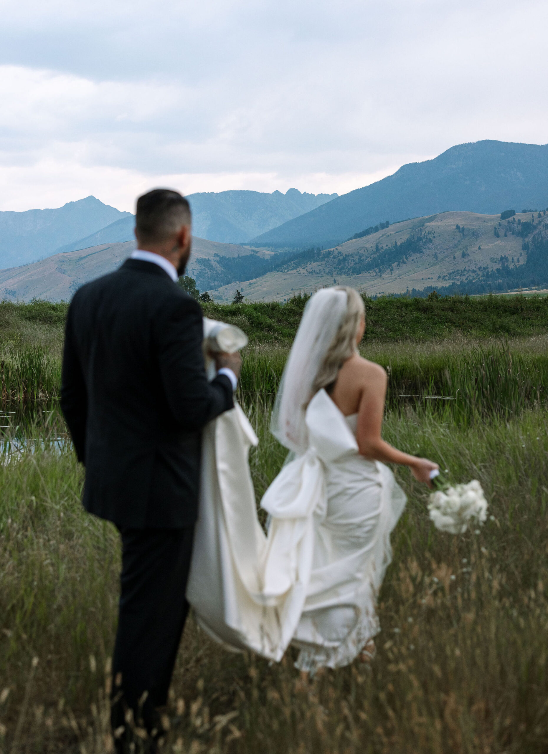 The bride and groom walk through a field of tall grass at Copper Rose Ranch Wedding Venue, with the Montana mountains in the background. The Montana Bride’s long veil flows behind her, and the scene is serenely captured by Haley J Photo, a Montana Wedding Venue Photographer.