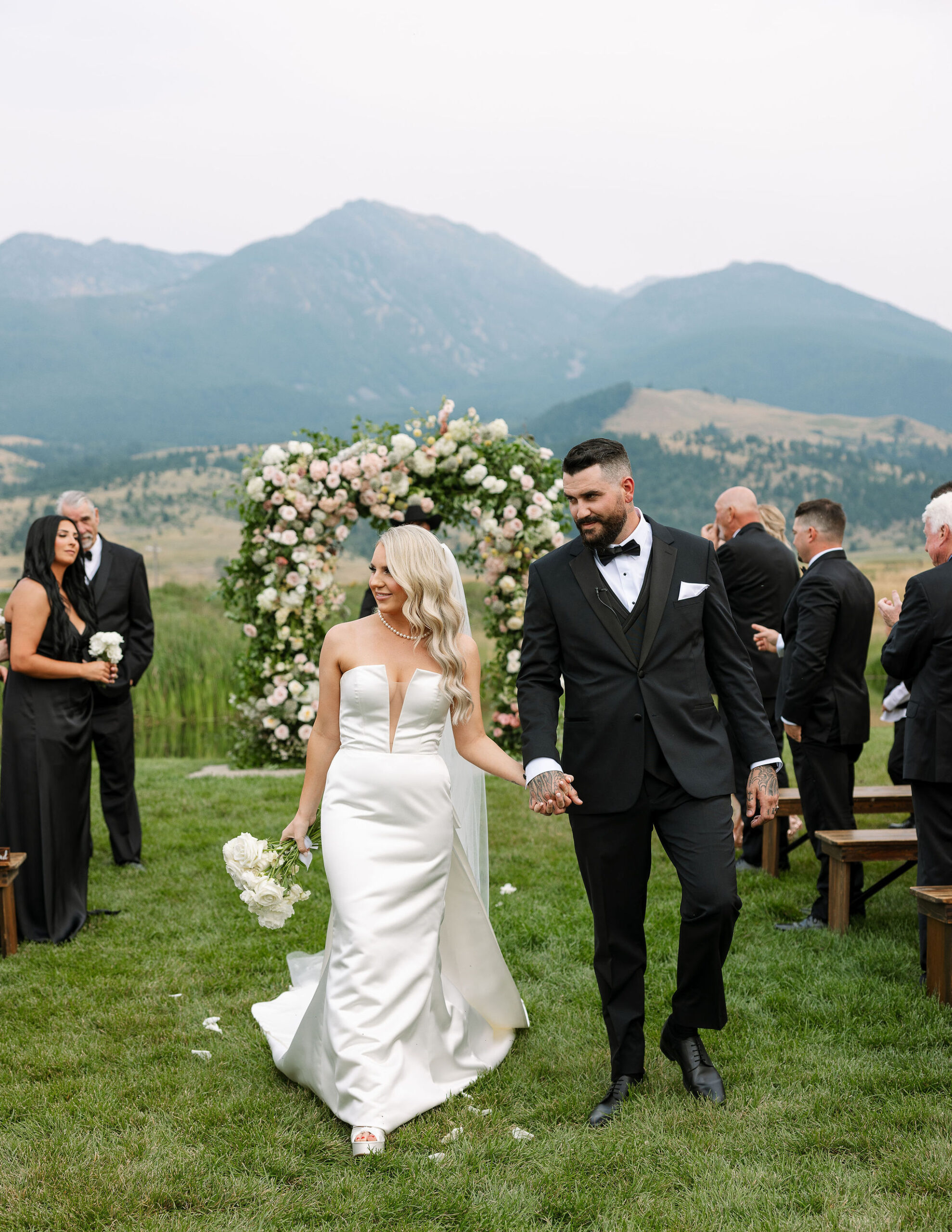 The bride and groom, hand in hand, walk down the aisle at Copper Rose Ranch Wedding Venue in Montana, with majestic mountains as the backdrop. The Montana Bride holds a bouquet of white flowers, and both are elegantly captured by Haley J Photo, a Montana Wedding Venue Photographer.