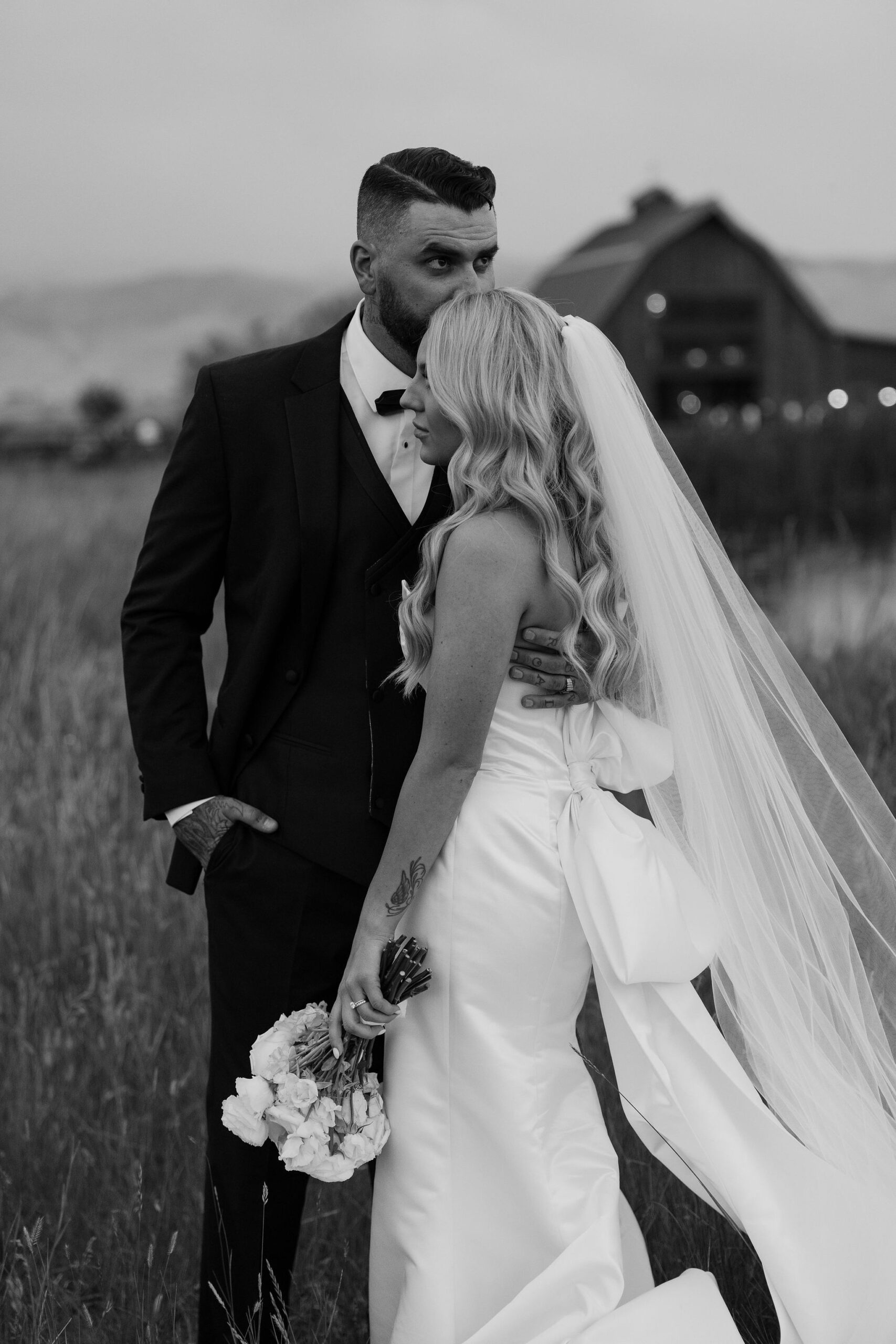 The groom kisses the Montana Bride on the forehead as they stand in a field at Copper Rose Ranch Wedding Venue. The black and white image highlights the intimate moment, beautifully captured by Haley J Photo, a Montana Wedding Photographer.