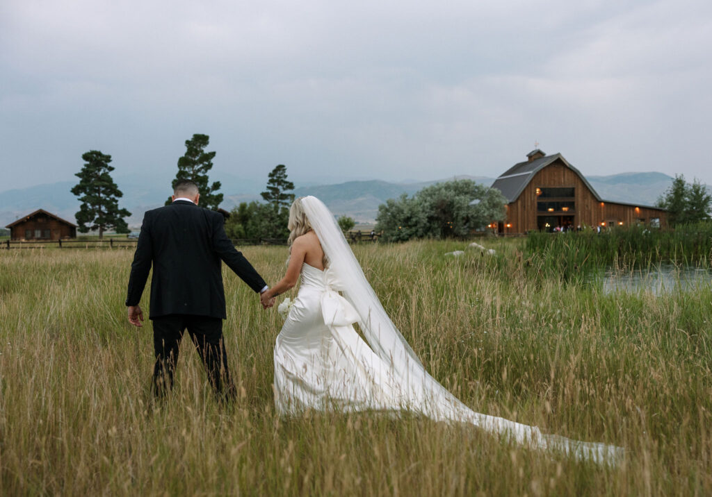 The bride and groom walking hand in hand through a field at Copper Rose Ranch Wedding Venue, with the venue's barn and Montana mountains in the background. Captured by Haley J Photo, a Montana Wedding Venue Photographer.