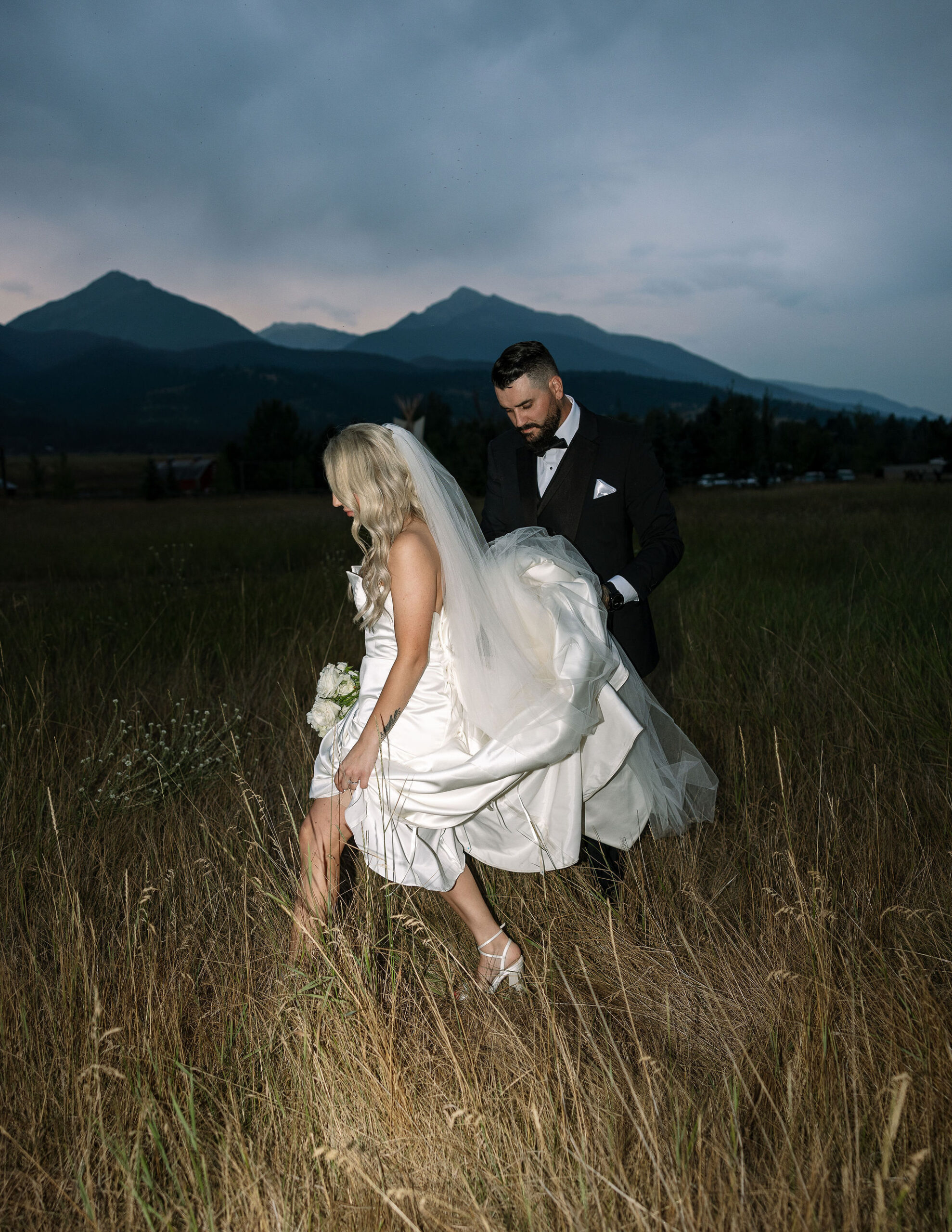 The bride and groom walk hand in hand through a field of tall grass at Copper Rose Ranch Wedding Venue in Montana, with mountains in the background. The intimate moment is beautifully captured by Haley J Photo, a Montana Wedding Venue Photographer.