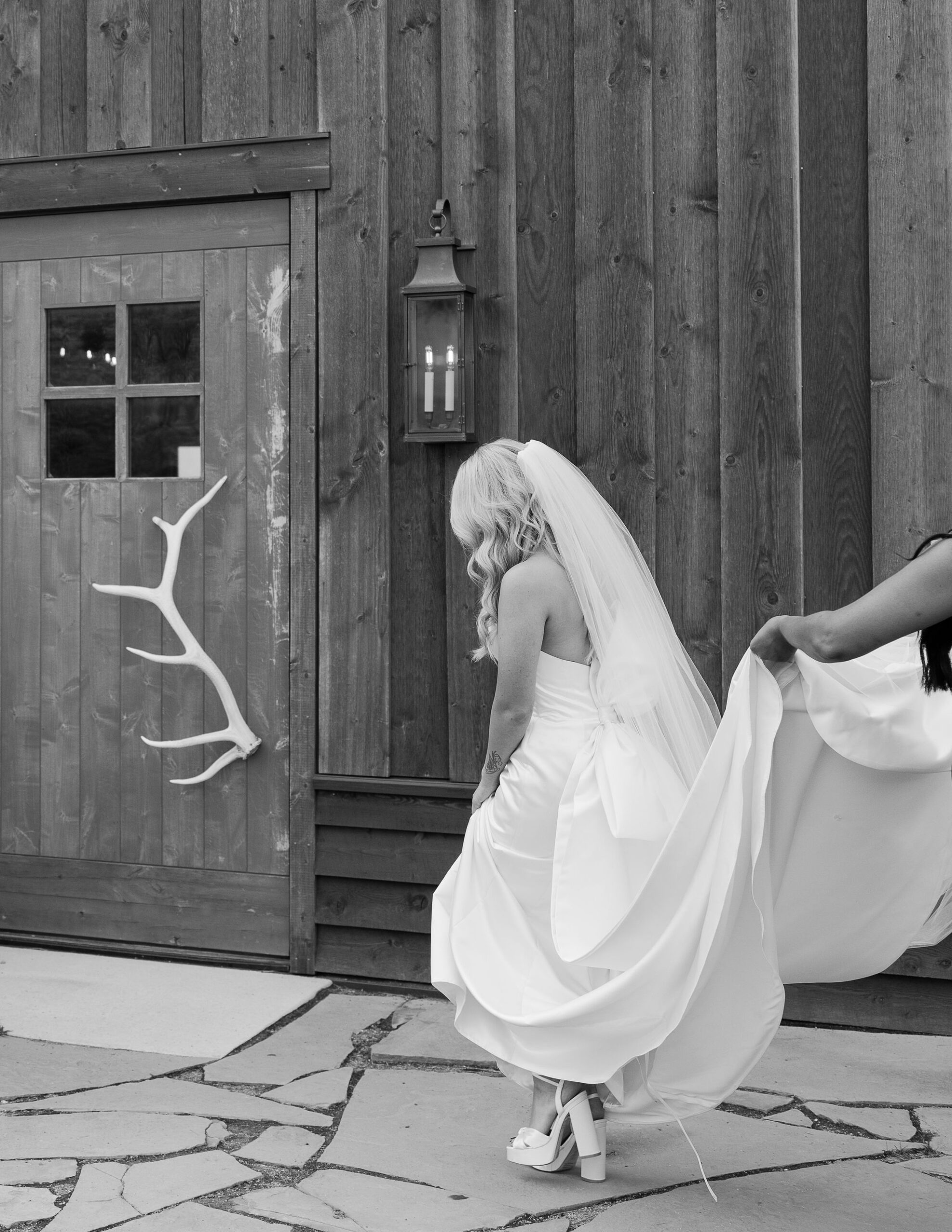 The bride, wearing a flowing white dress, walks towards the entrance of a rustic barn at Copper Rose Ranch Wedding Venue. The black and white photo emphasizes the contrast between the dress and the wooden structure, captured by Montana Wedding Photographer Haley J Photo.