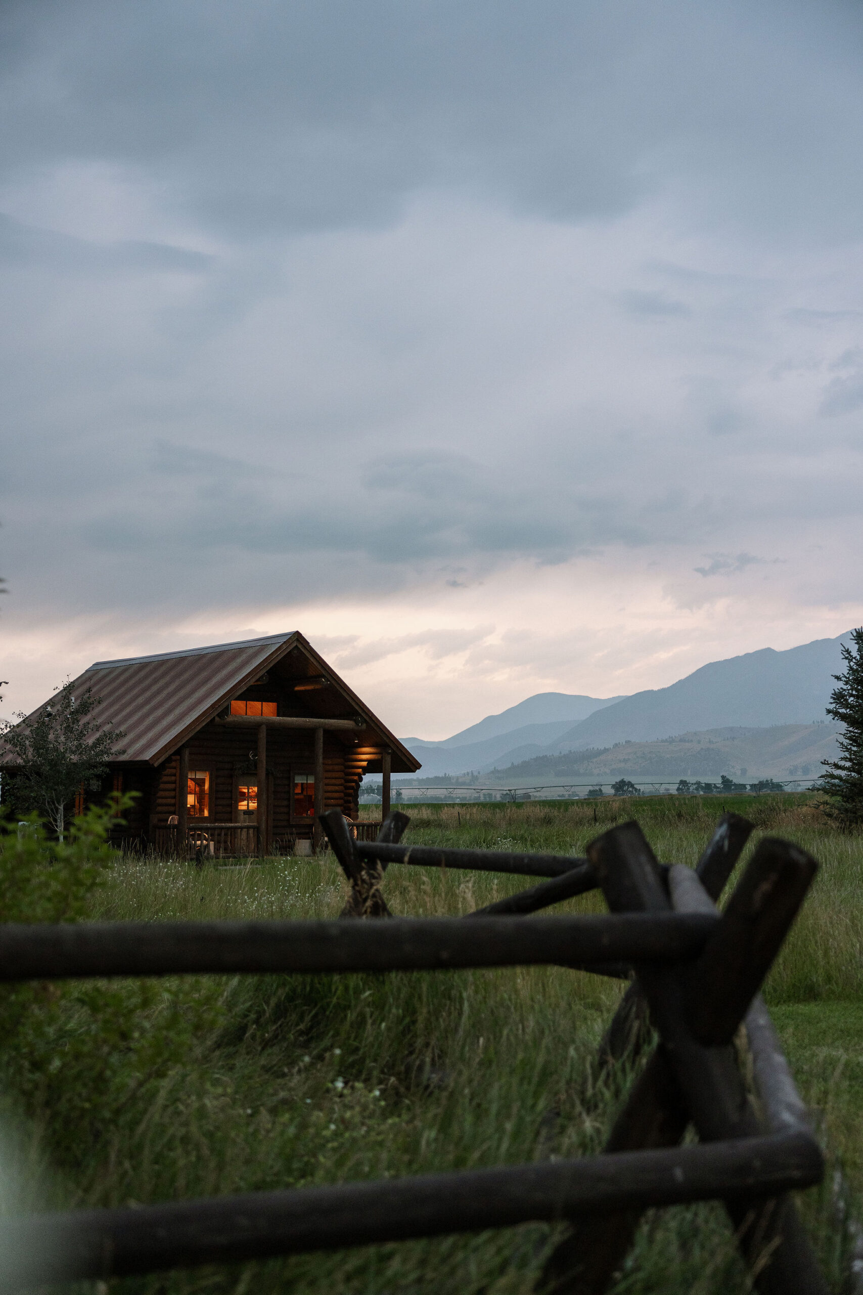 A serene view of a cabin at Copper Rose Ranch Wedding Venue in Montana, surrounded by lush greenery and mountains in the distance. The evening light adds a peaceful ambiance, captured by Montana Wedding Venue Photographer Haley J Photo.