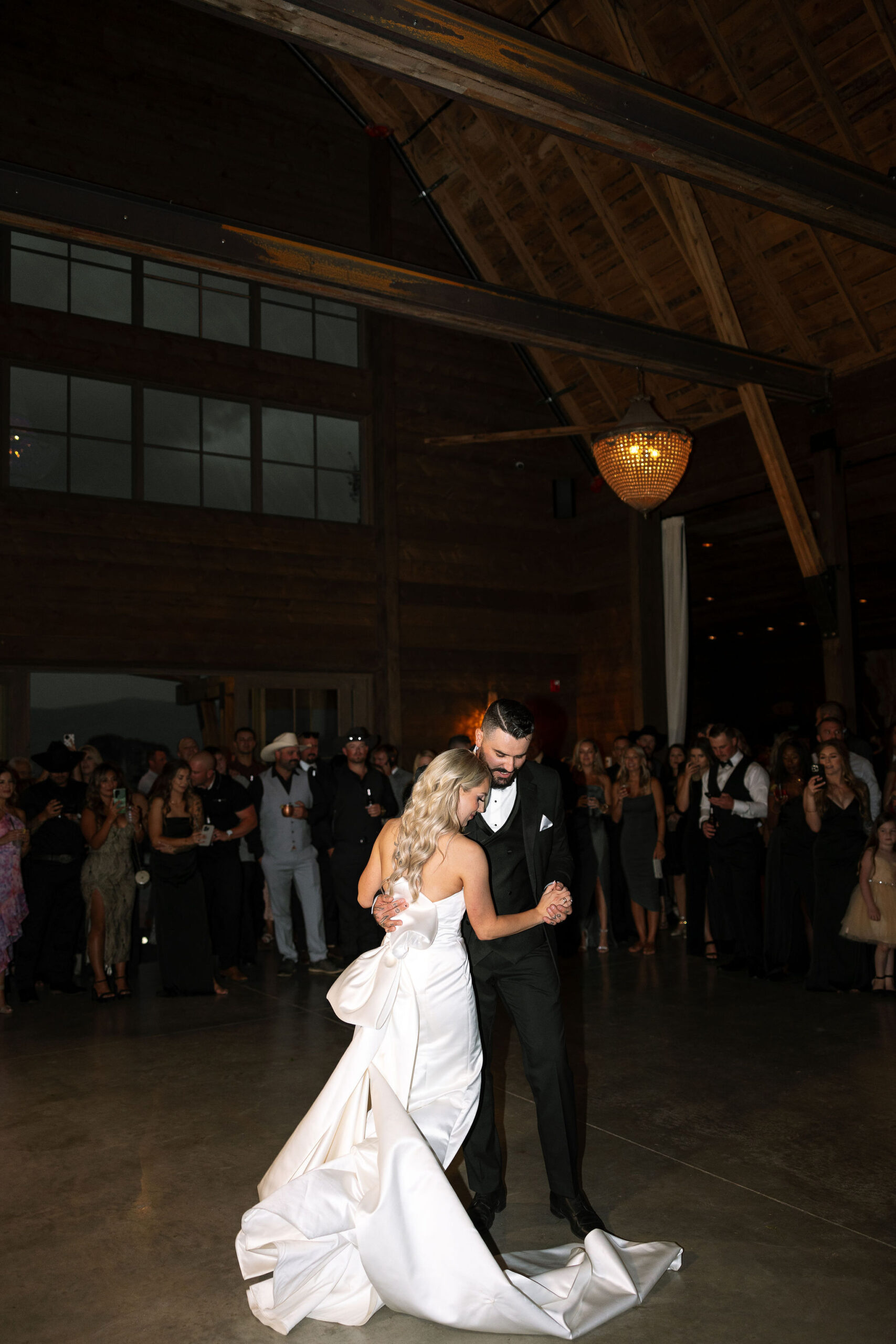 The bride and groom share their first dance inside the rustic barn at Copper Rose Ranch Wedding Venue. The warm wooden interior and chandeliers create an elegant setting, captured by Montana Wedding Venue Photographer Haley J Photo.